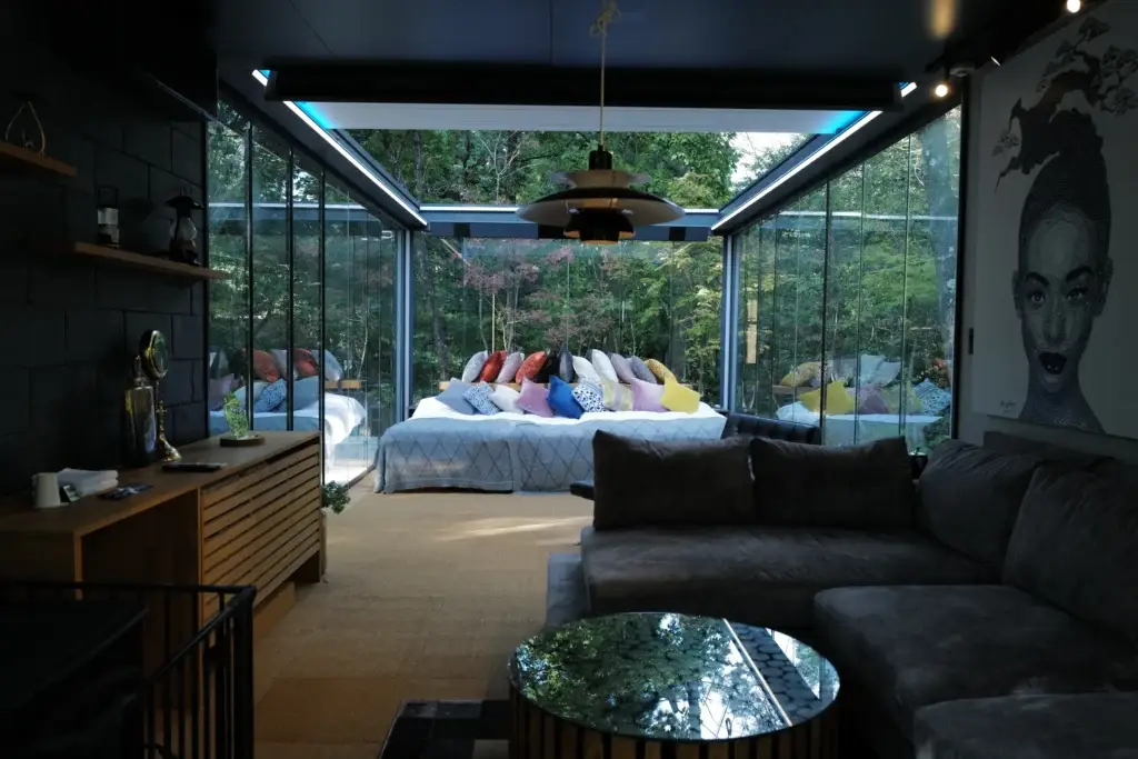 Interior of Hikari no Hako room showing bed with colorful pillows, sofa, and modern art through glass walls overlooking the forest.
