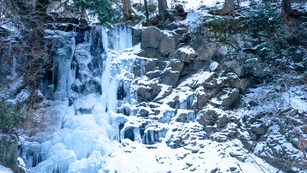 A wide view of the frozen Haha no Shirataki waterfall, showing the intricate patterns of ice clinging to the rock face.