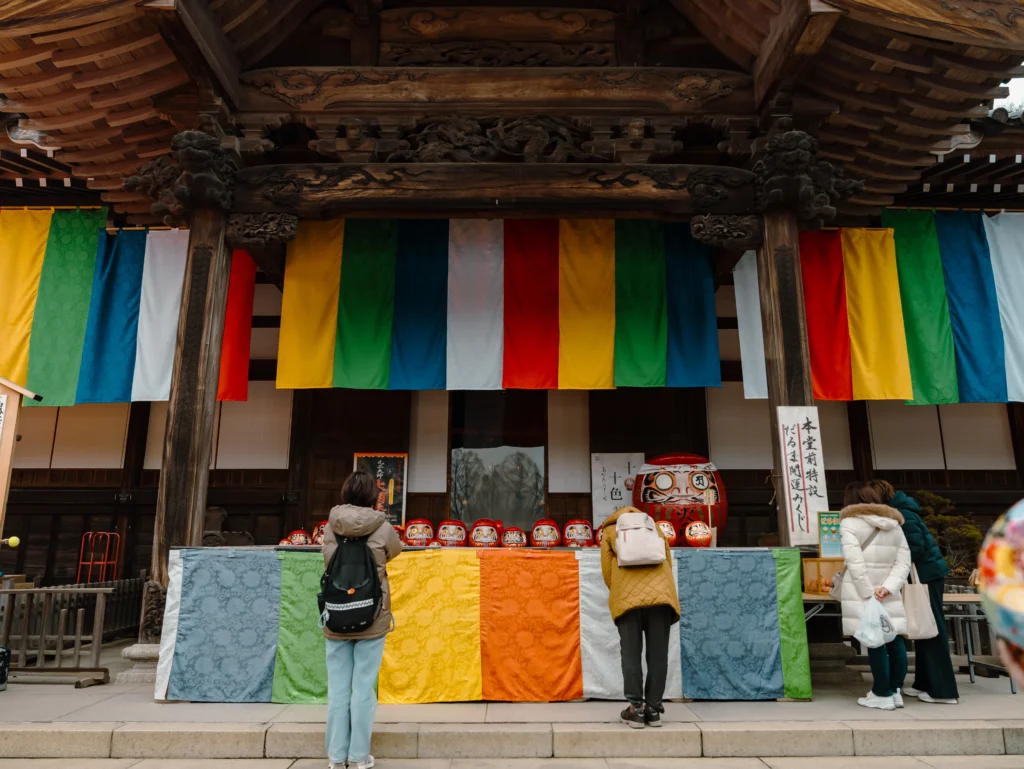 Visitors praying in front of the main hall of Jindaiji Temple, decorated with colorful Buddhist flags for the Daruma Ichi.