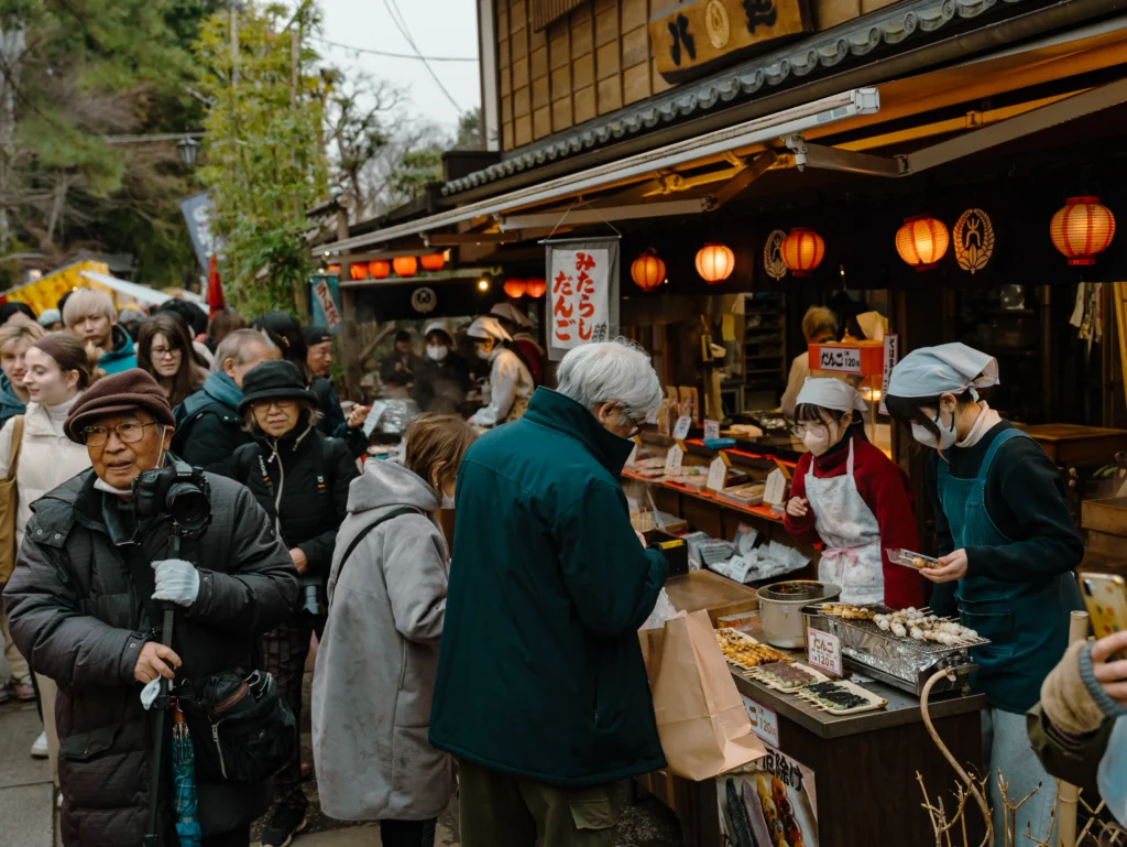 A lively crowd enjoys street food from a dango stall at the bustling Jindaiji Daruma Ichi.