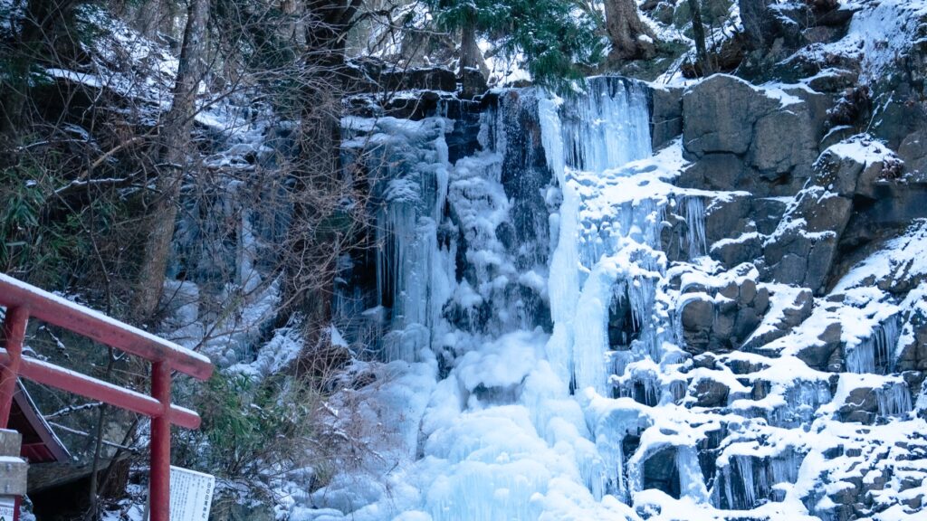 Haha no Shirataki waterfall frozen into thick blue-white columns of ice against dark volcanic rock, with snow on the ledges and a small vermilion torii and wooden shrine building standing at the left edge of the ravine.