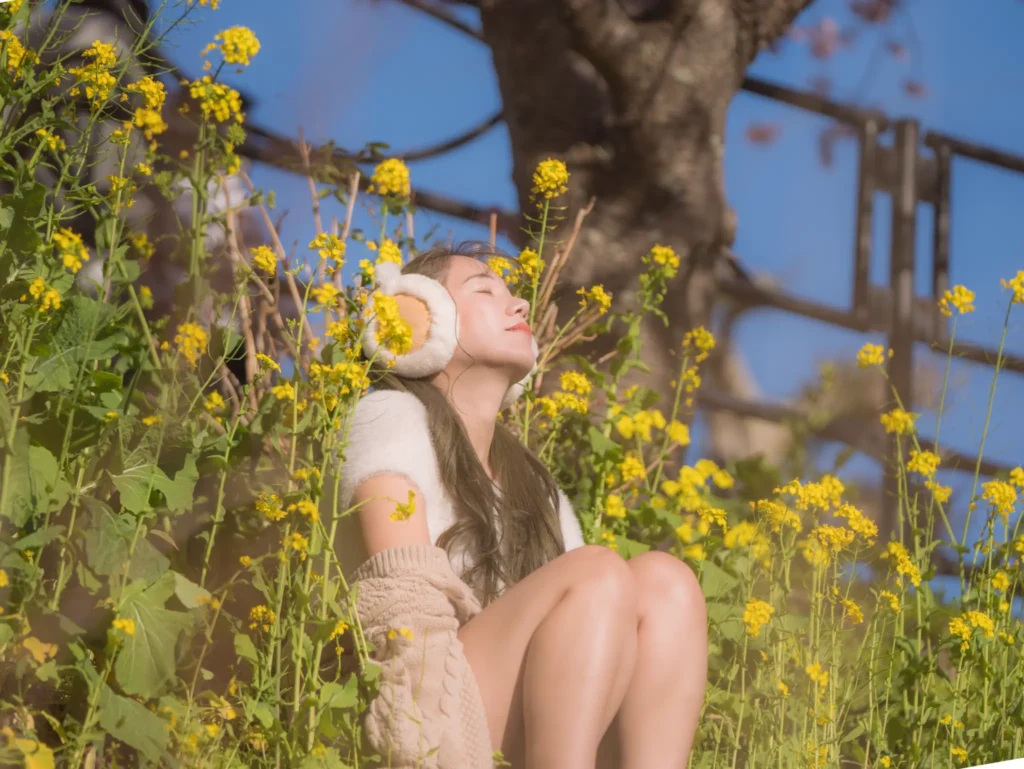 A woman wearing earmuffs relaxes among yellow nanohana (rapeseed) flowers with Kawazu cherry blossoms visible in the background under a clear blue sky.
