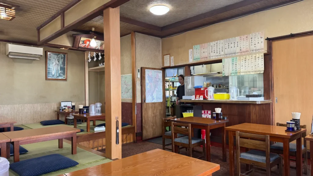 Inside Gensuke restaurant showing traditional tatami seating on the left, table seating on the right, and an open kitchen in the center where staff prepare meals, with handwritten menu boards lining the walls.