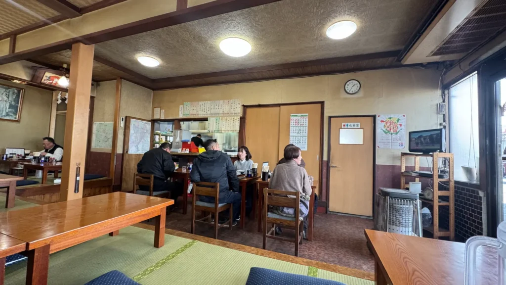 The lively interior of Gensuke during lunchtime, with local customers enjoying their meals at wooden tables, tatami seating visible in the background, and a homey atmosphere with wall clock and calendar.