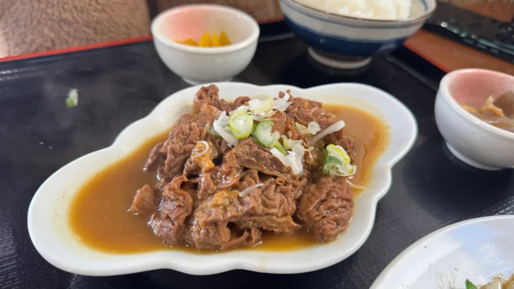 Close-up of Motsu Ni (simmered pork offal) at Gensuke, featuring tender pieces of pork intestines braised in a rich miso-based sauce, topped with chopped green onions, served in a decorative white dish.