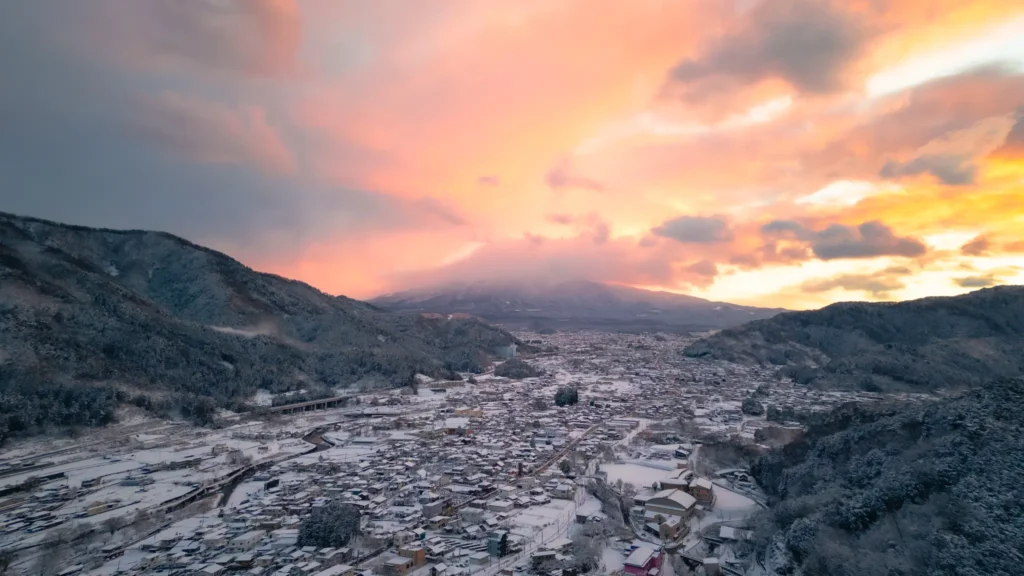 A dramatic sunset with orange and pink clouds completely hiding Mount Fuji, seen from above a snow-covered town.