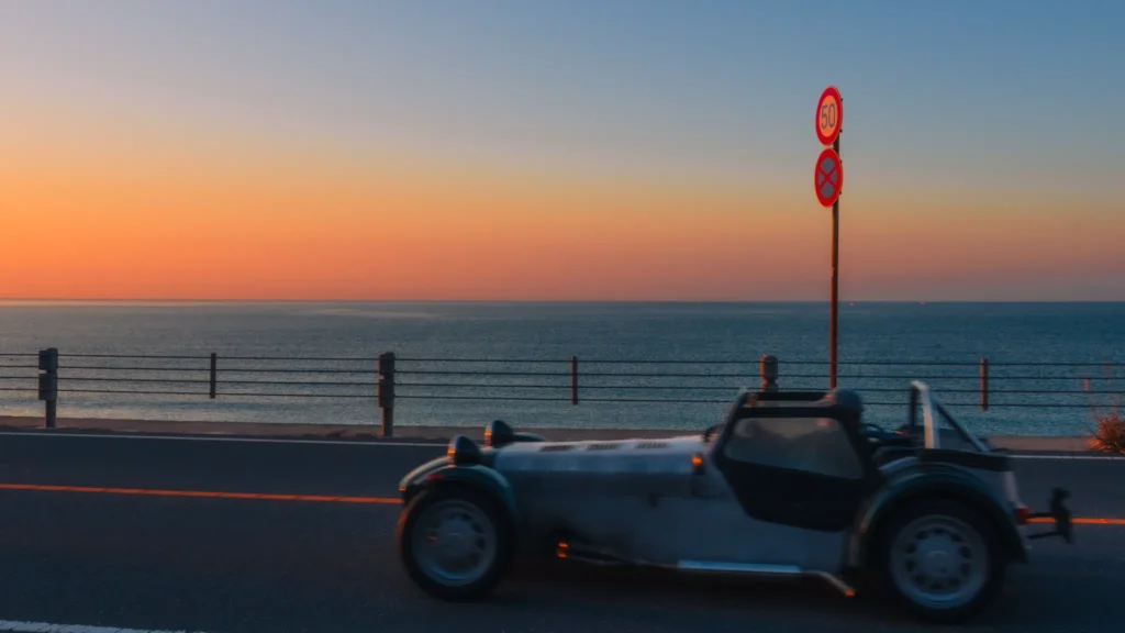 A classic silver convertible car on Route 134 at sunrise, with the calm blue sea of Shichirigahama in the background.