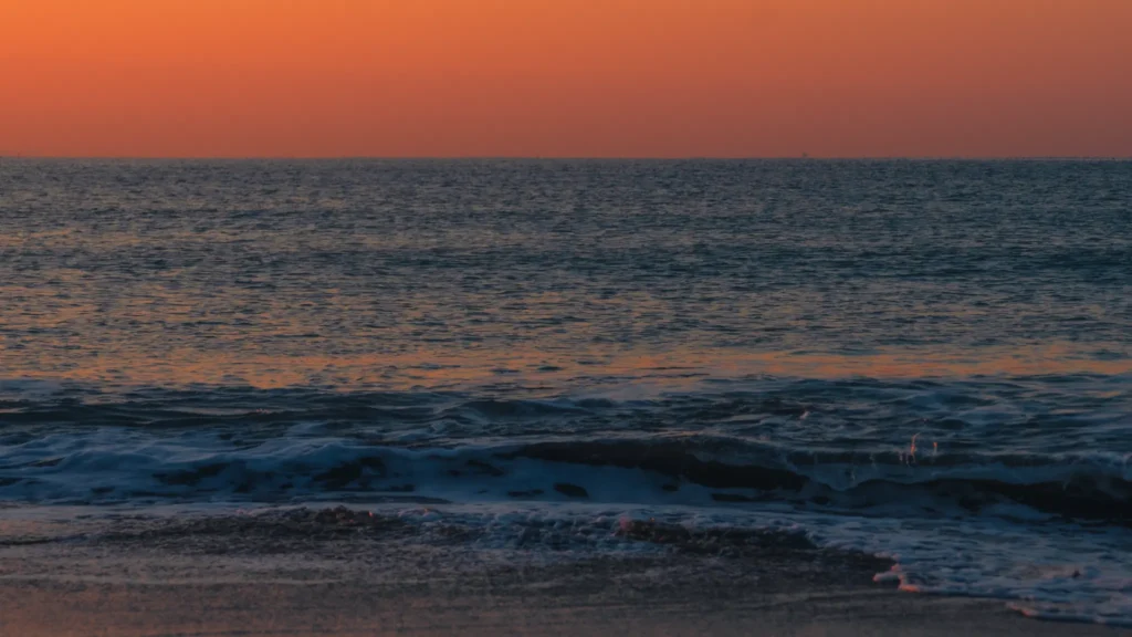 A close-up of gentle waves on the dark sand of Shichirigahama beach, reflecting the orange glow of the sunrise.