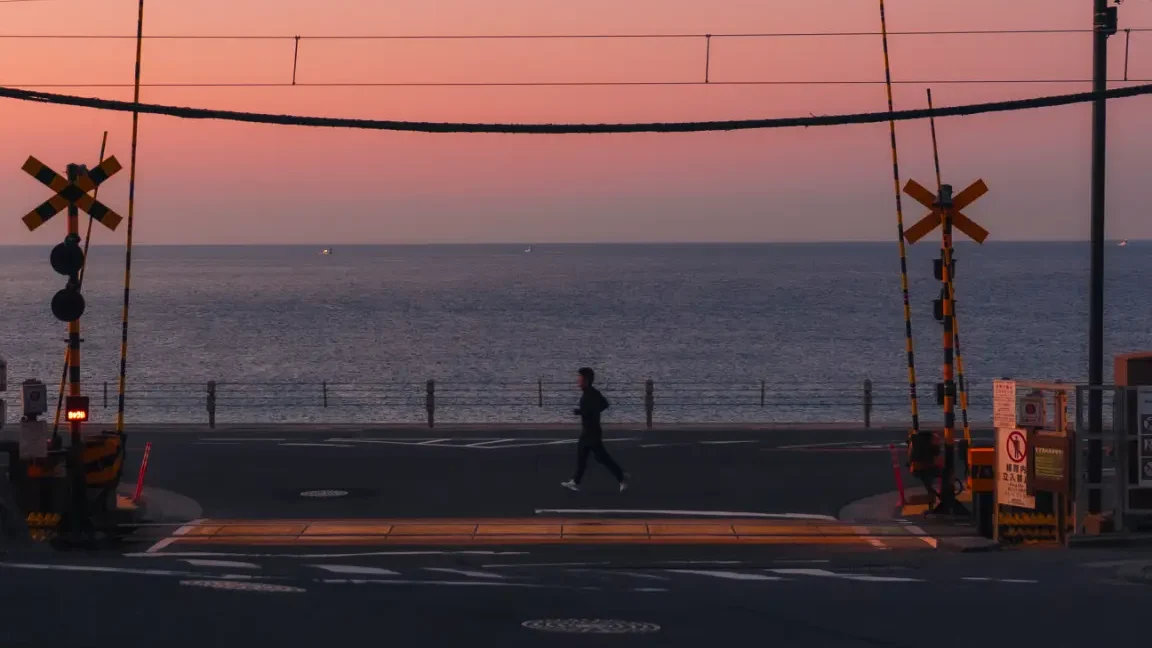 A lone person crosses the Slam Dunk railway tracks at sunrise, silhouetted against a pink and orange sky over the ocean.