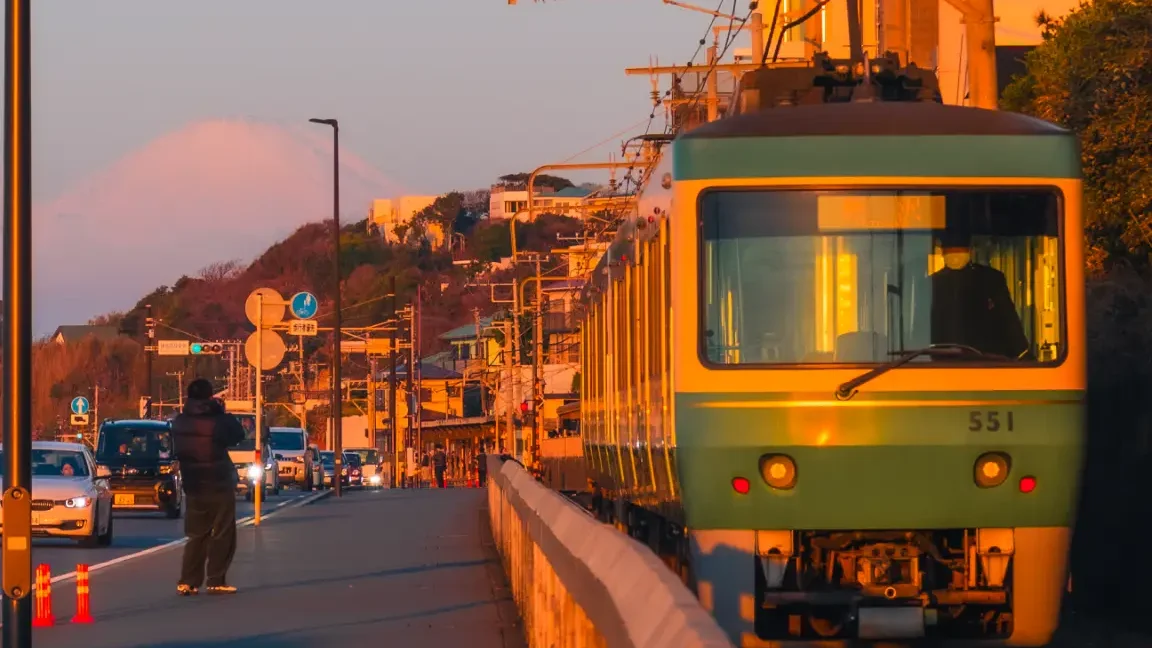 The green and yellow Enoden train approaches Kamakura-koko-mae station during the golden hour, with a silhouette of Mt. Fuji visible in the background.