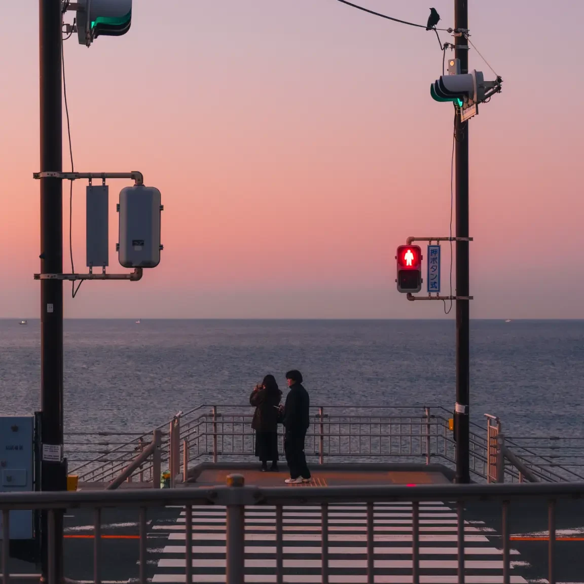 Two people stand silhouetted against a pink and purple sunrise, looking out at the sea from the traffic light near the Slam Dunk crossing.