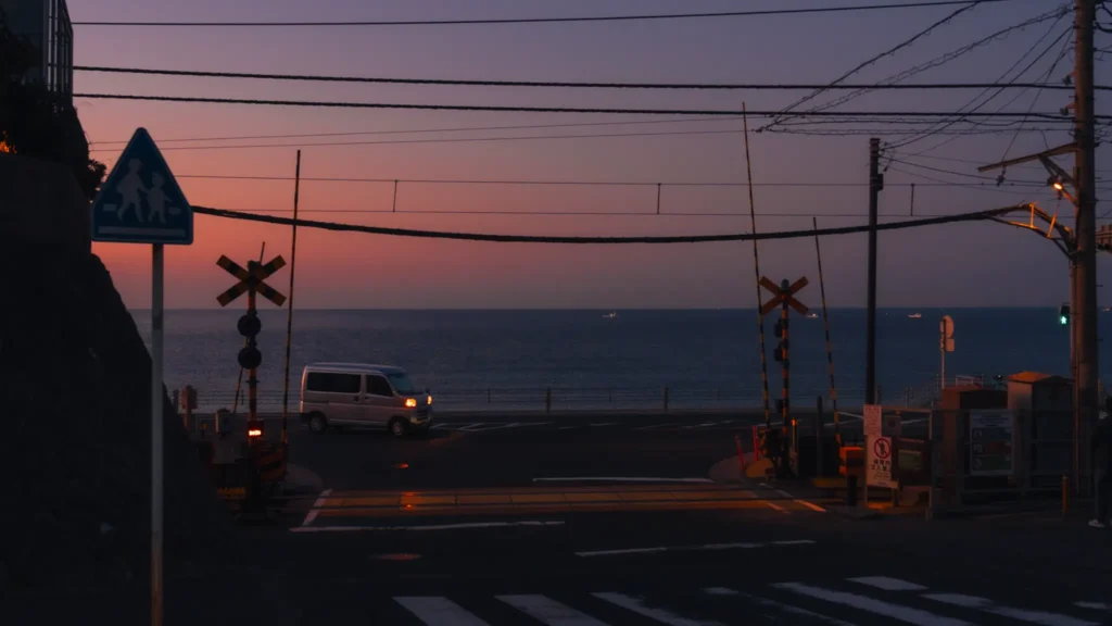 The Kamakura Koko-mae crossing viewed from the hill at dawn, with a van passing through and the Shonan coast glowing pink and orange in the background.