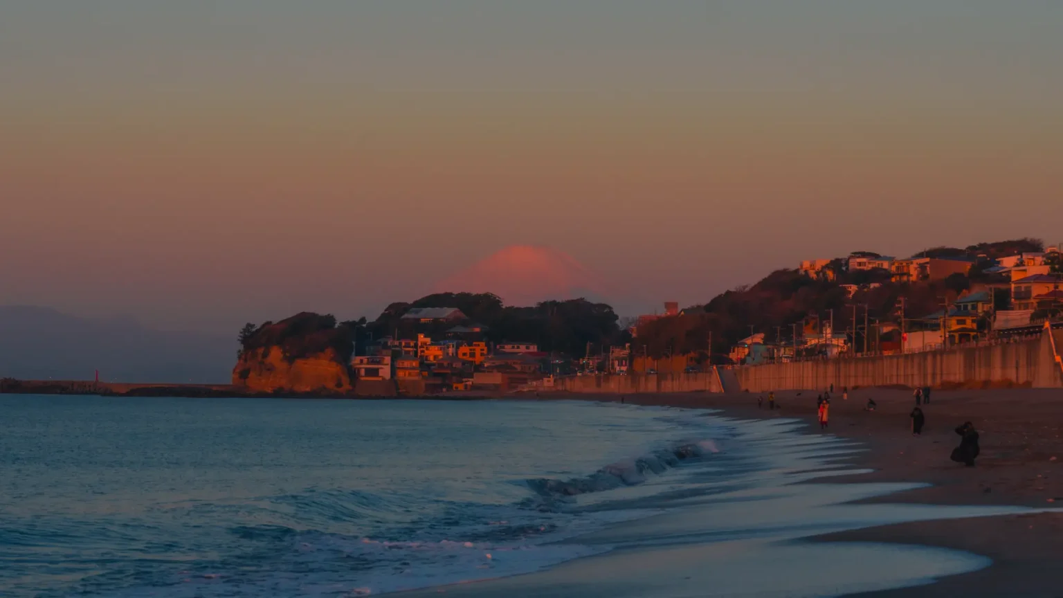 The silhouette of Mt. Fuji glows pink behind Inamuragasaki cape, viewed from the shores of Shichirigahama beach at sunset.