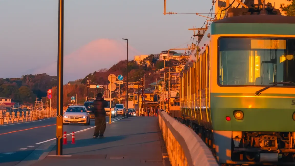 The green and yellow Enoden train travels along the coast during golden hour, with the silhouette of Mt. Fuji visible in the background.