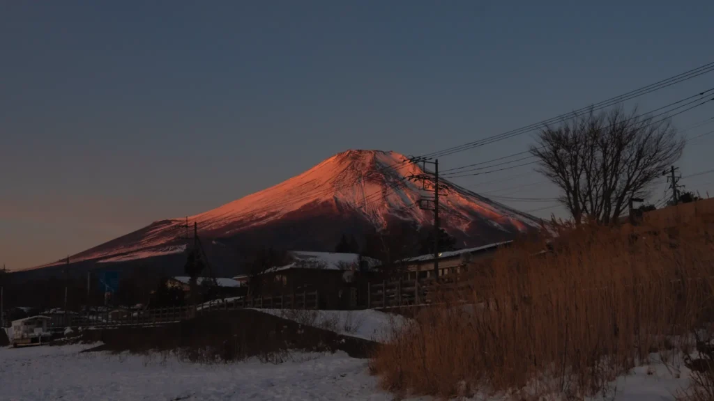 Mt. Fuji illuminated by the morning sun, glowing red against a dark blue sky, seen from the snowy shores of Lake Yamanakako.