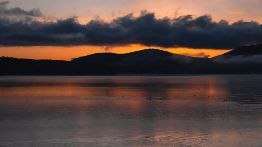 The calm, dark waters of Lake Yamanakako just before dawn, with the orange glow of sunrise reflecting on the surface.