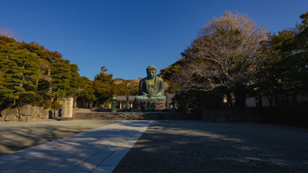 A wide shot of the Great Buddha statue in the temple grounds, with the 'National Treasure Great Buddha of Kamakura' stone marker in the foreground.
