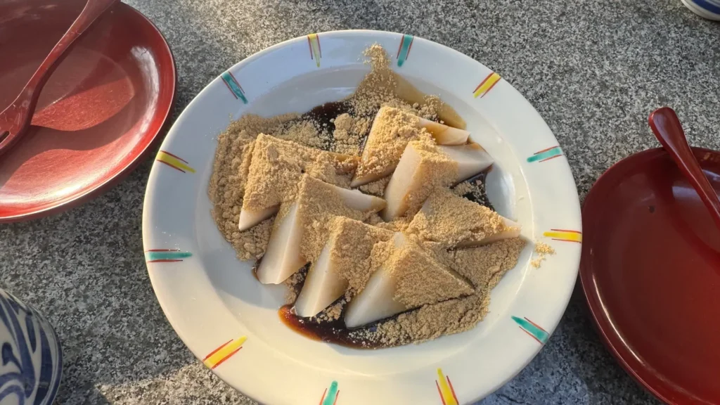Kuzumochi (arrowroot starch cake) dessert at Suzume no Oyado, featuring triangular slices of translucent jelly-like mochi generously dusted with kinako (roasted soybean flour) and drizzled with kuromitsu (black sugar syrup).