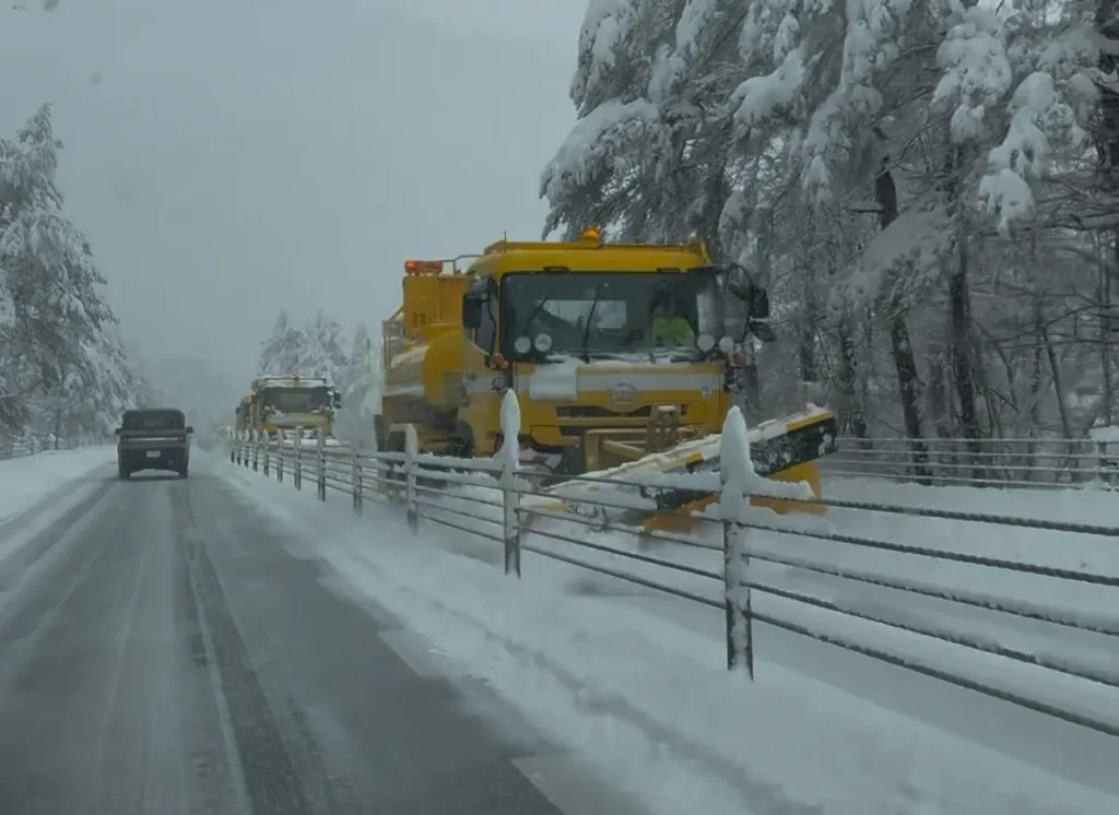 A yellow snow plow truck clearing a road covered in deep snow, highlighting the winter driving conditions near Mt. Fuji.
