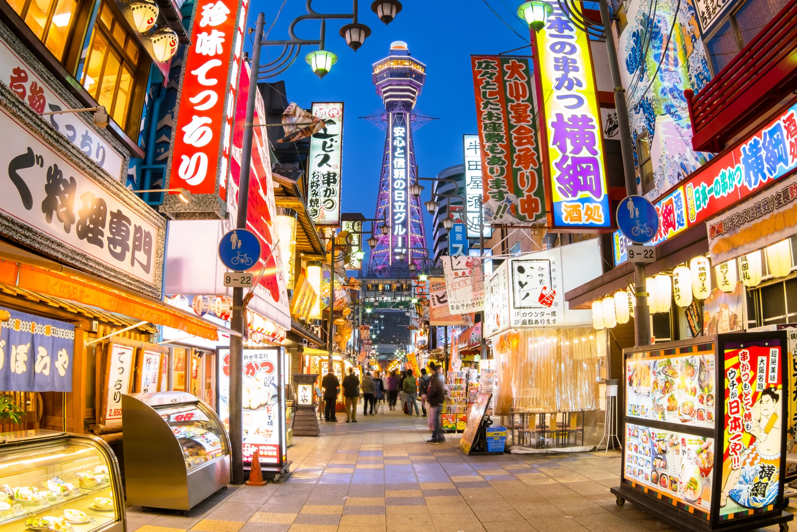 A bustling street in Osaka at night, with bright neon signs and the Tsutenkaku Tower in the background.