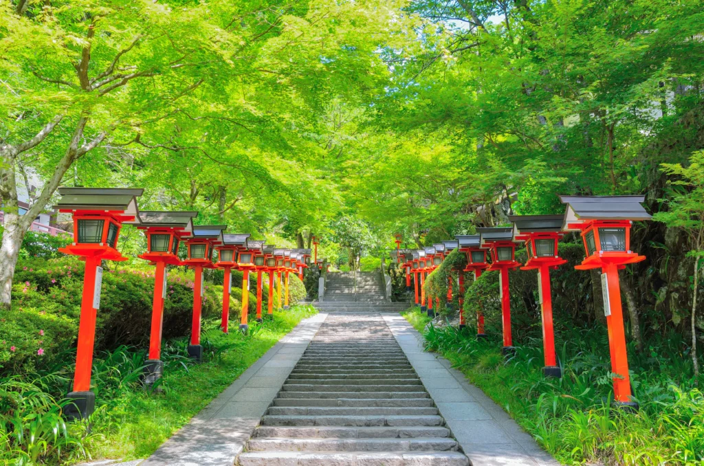 A stone staircase lined with red lanterns leads up to a temple entrance, surrounded by lush green trees.