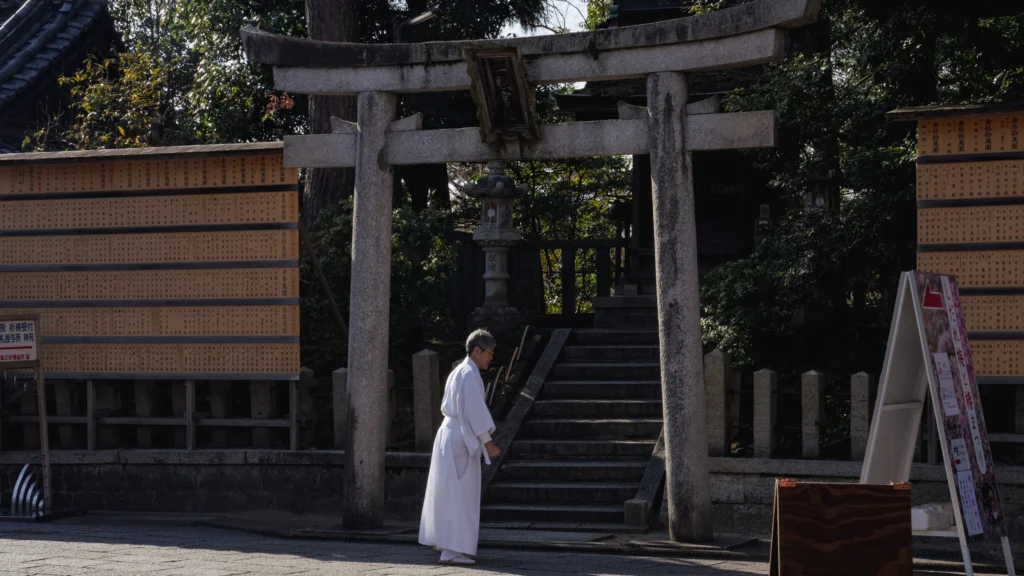A Shinto priest in white robes walking through a stone torii gate at Jonangu Shrine, with donor name boards lining both sides of the path.