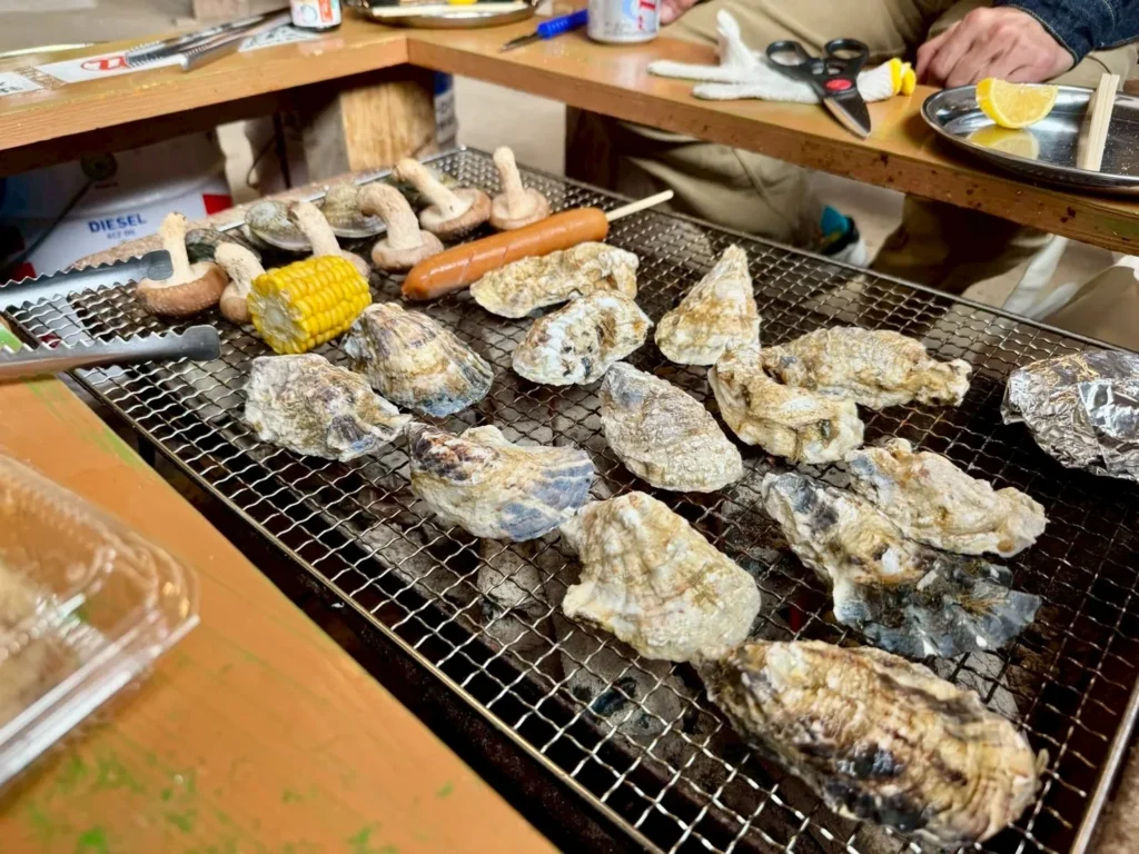 A variety of foods grilling over a charcoal fire, including oysters, shiitake mushrooms, corn on the cob, and a sausage.