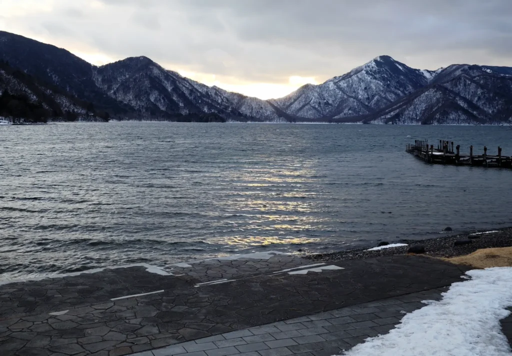 A view of Lake Chuzenji in winter, with a wooden pier and snow-covered mountains in the background.