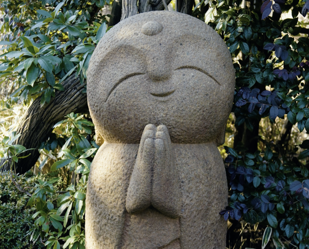 A large smiling stone Jizo statue with hands in prayer at Hasedera temple in Kamakura, surrounded by green foliage.