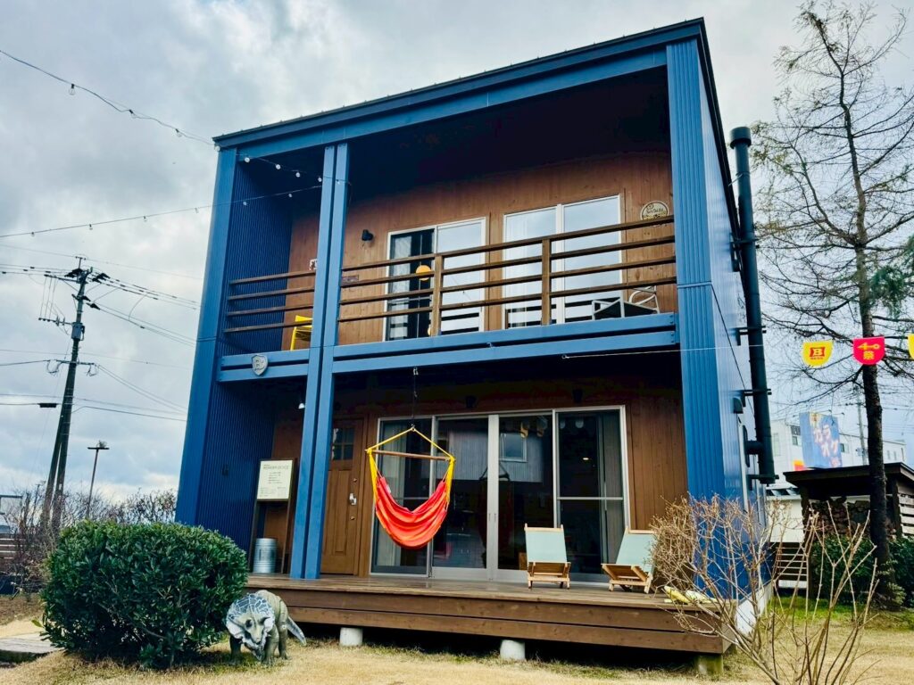 A blue WONDER DEVICE model house at BESS Kumamoto with a spacious wooden deck and a hammock, inviting visitors to relax.