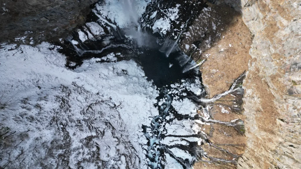 Bird's-eye drone view looking straight down at the base of Kegon Falls in winter, showing the dark pool and surrounding ice and snow.