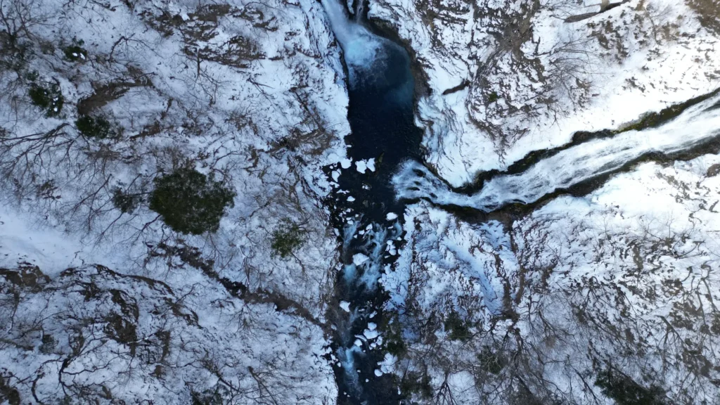 Drone view from above the top of Kegon Falls in winter, showing the water flowing through a snow-covered gorge before plunging over the cliff edge.