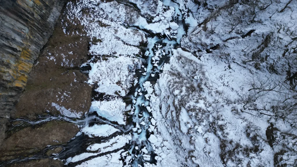 Aerial view of the frozen stream below Kegon Falls in winter, with ice chunks and snow-covered rocks along the gorge.