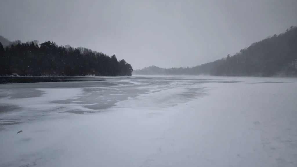 The frozen surface of Lake Chuzenji covered in snow, with misty mountains in the background during a winter snowfall in Nikko.