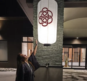 A person in a traditional yukata reaches up to a large, glowing white lantern at the entrance of Kamenoi Hotel Oku-Nikko at night, with snow on the ground.