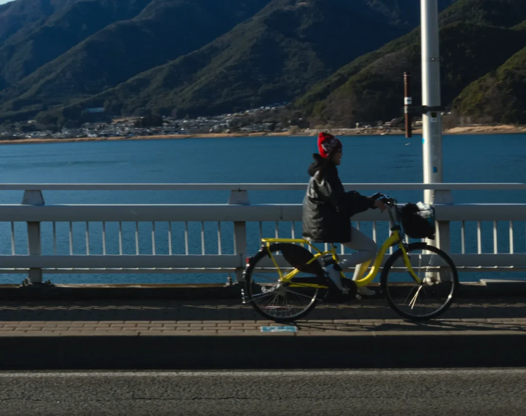 A person wearing a red beanie cycles across Kawaguchiko Ohashi Bridge on a bright yellow rental bike, with the blue lake and mountains in the background.