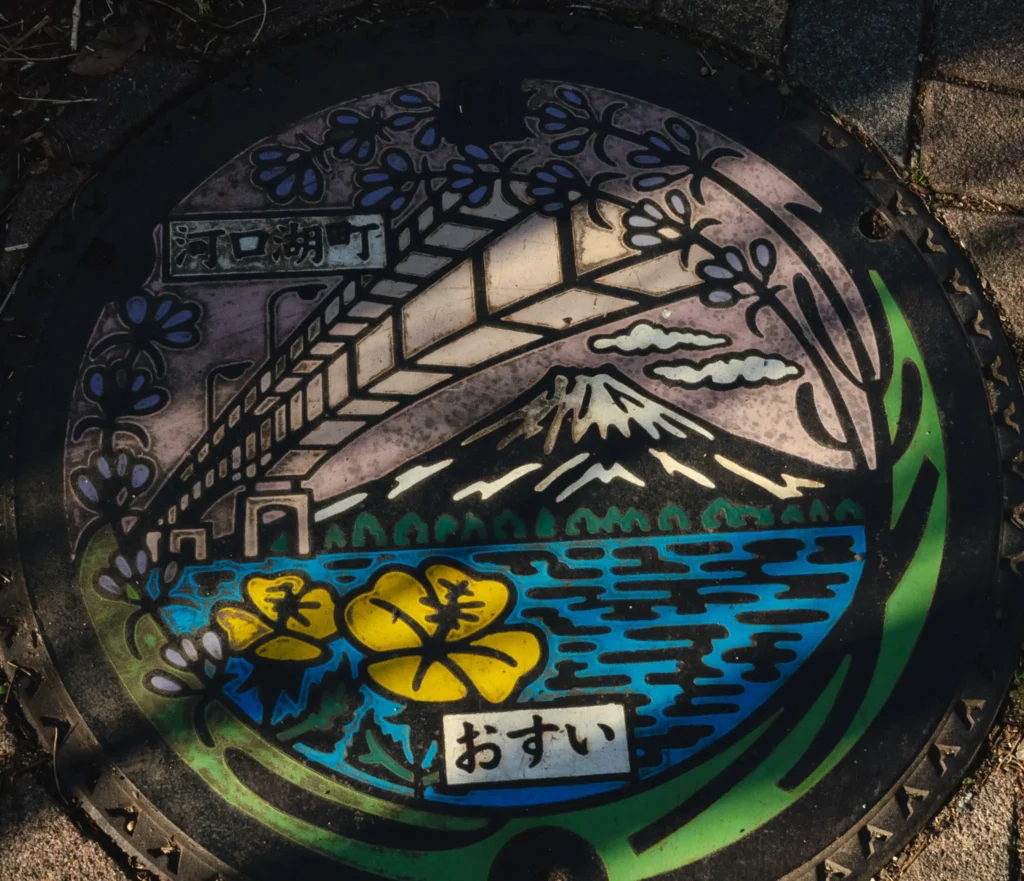 A colorful manhole cover in Fujikawaguchiko, featuring a design of the Kawaguchiko Ohashi Bridge, Mt. Fuji, and yellow hibiscus flowers.