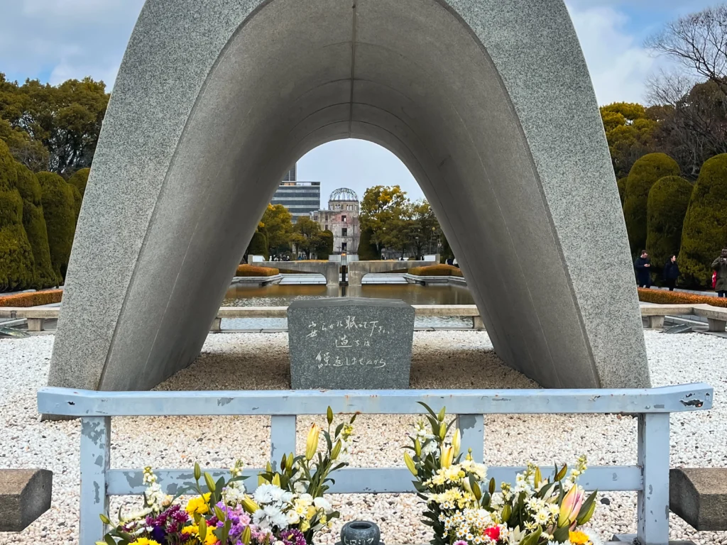 The Cenotaph for Atomic Bomb Victims with flower offerings, the Peace Flame and Dome visible through the arch

