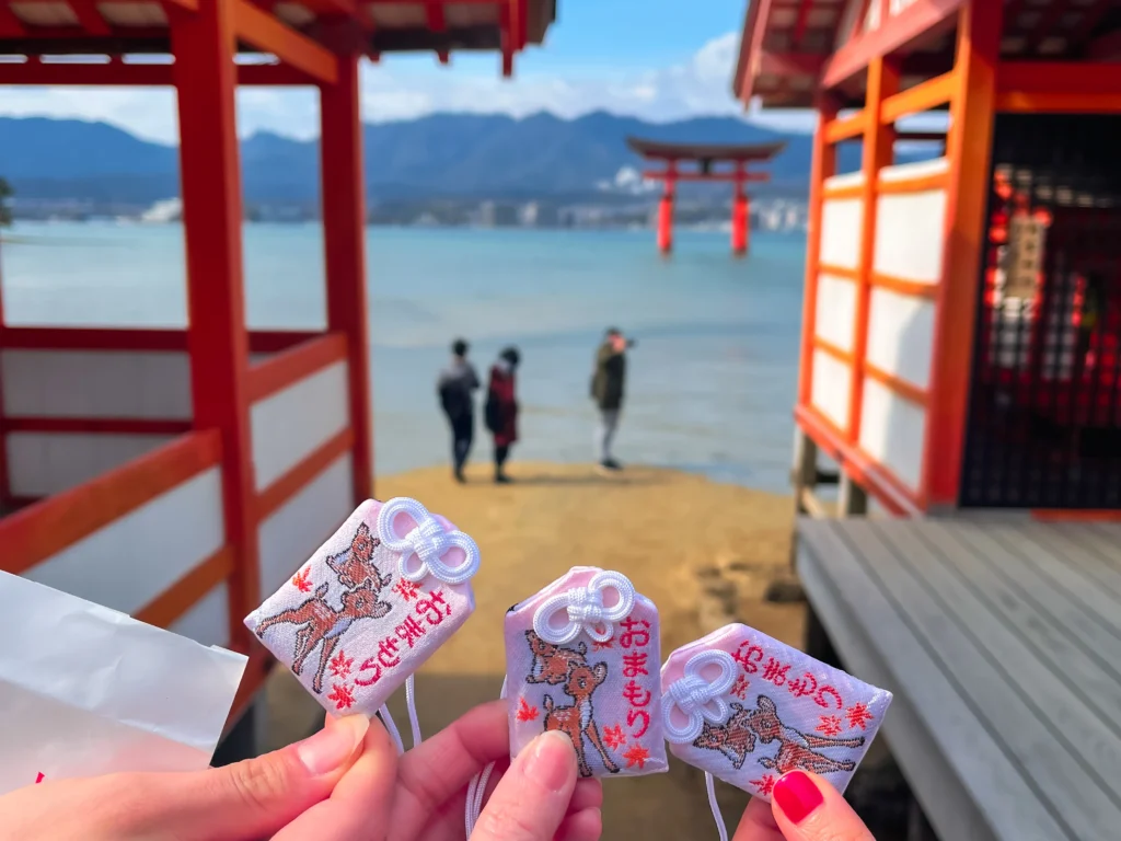 Three Itsukushima Shrine omamori charms held up with the floating torii gate behind