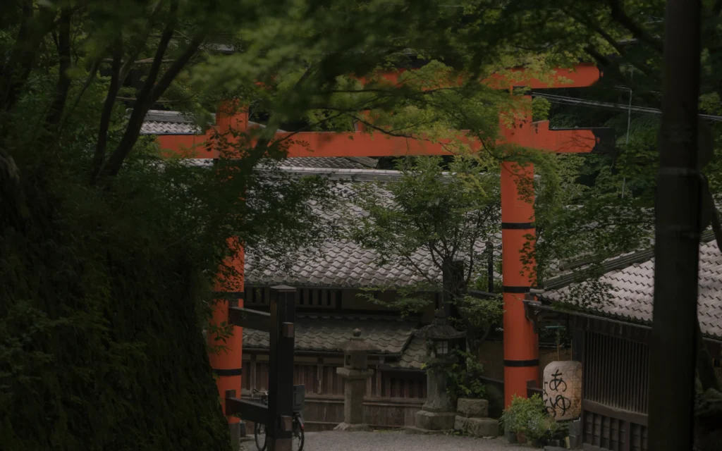 The vermilion first torii gate of Atago Shrine framed between trees, with traditional roof tiles and a stone lantern visible in the foreground