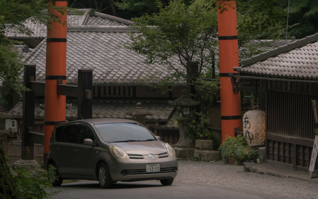 Two vermilion torii pillars flanking the road with traditional buildings, a stone lantern, and a compact car passing underneath