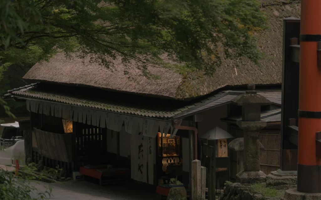 Thatched roof traditional tea house with dark wooden walls, noren curtains, and a stone lantern, nestled into the green forest