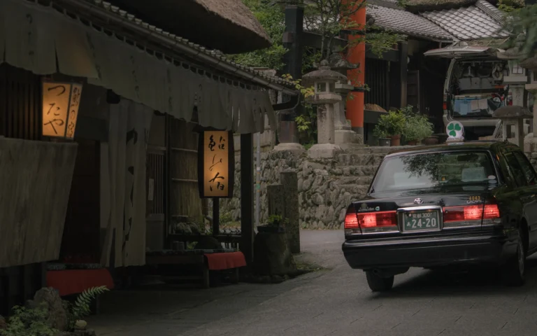 A quiet lane in Kyoto's preserved Saga-Toriimoto district at dusk: a thatched teahouse with paper lanterns, stone lanterns and a vermilion torii gate, with a classic Japanese taxi passing and no crowds in sight