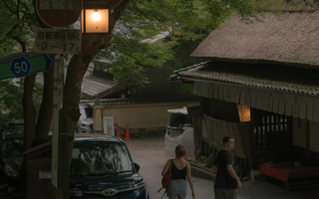 Street view with a hanging street lamp, a road sign showing "50 km/h zone 9-17", and two visitors walking past a thatched-roof building in late afternoon light