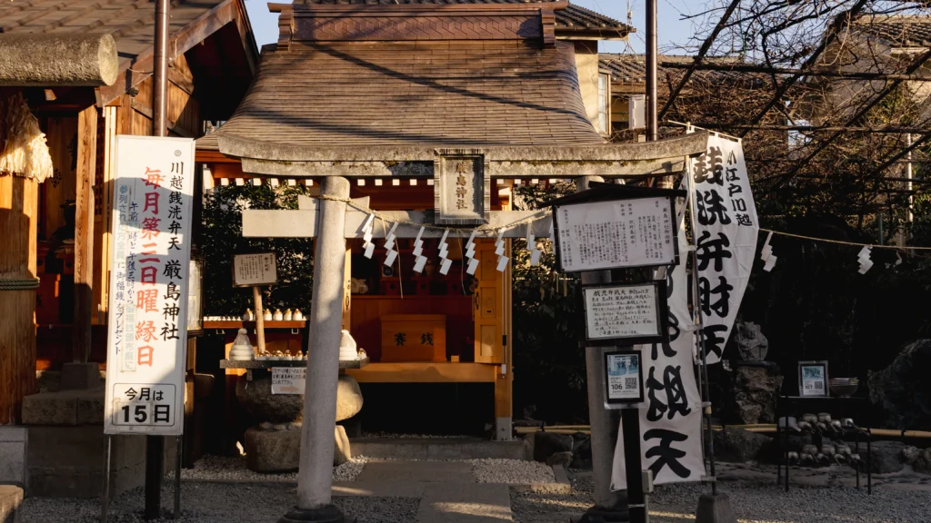 Zeniarai Benten money-washing shrine at Kawagoe Kumano with Itsukushima Shrine torii gate, banners reading zeni-arai benten, and the monthly ritual schedule sign