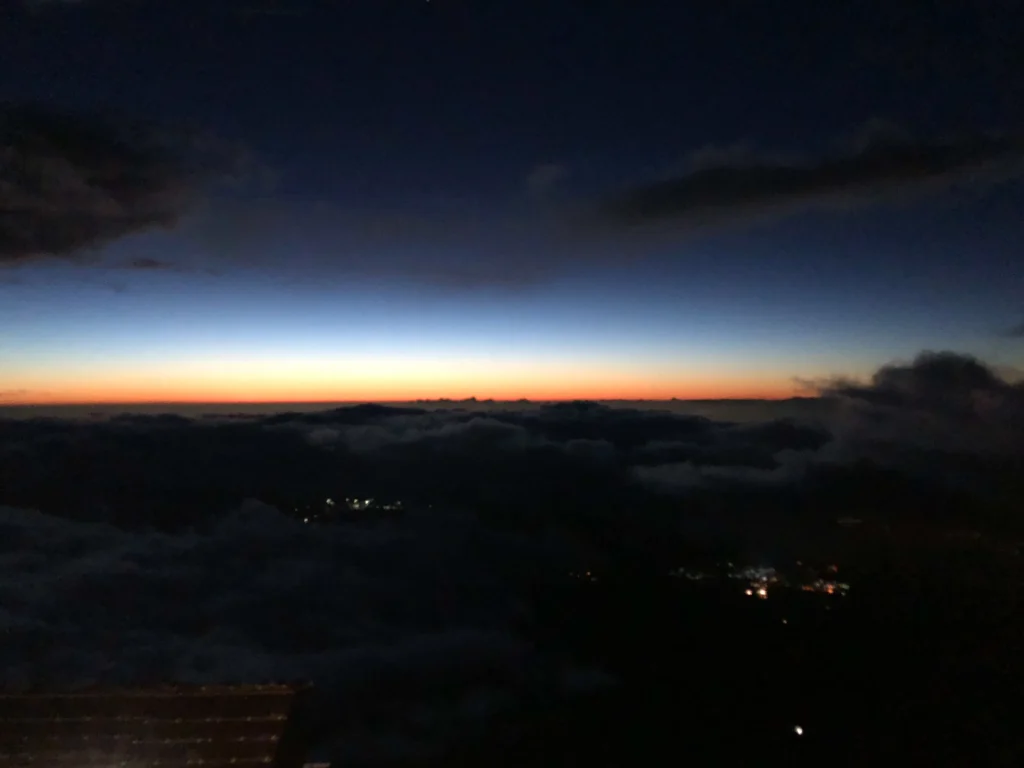 First orange glow on the horizon beneath deep blue sky, with clouds below