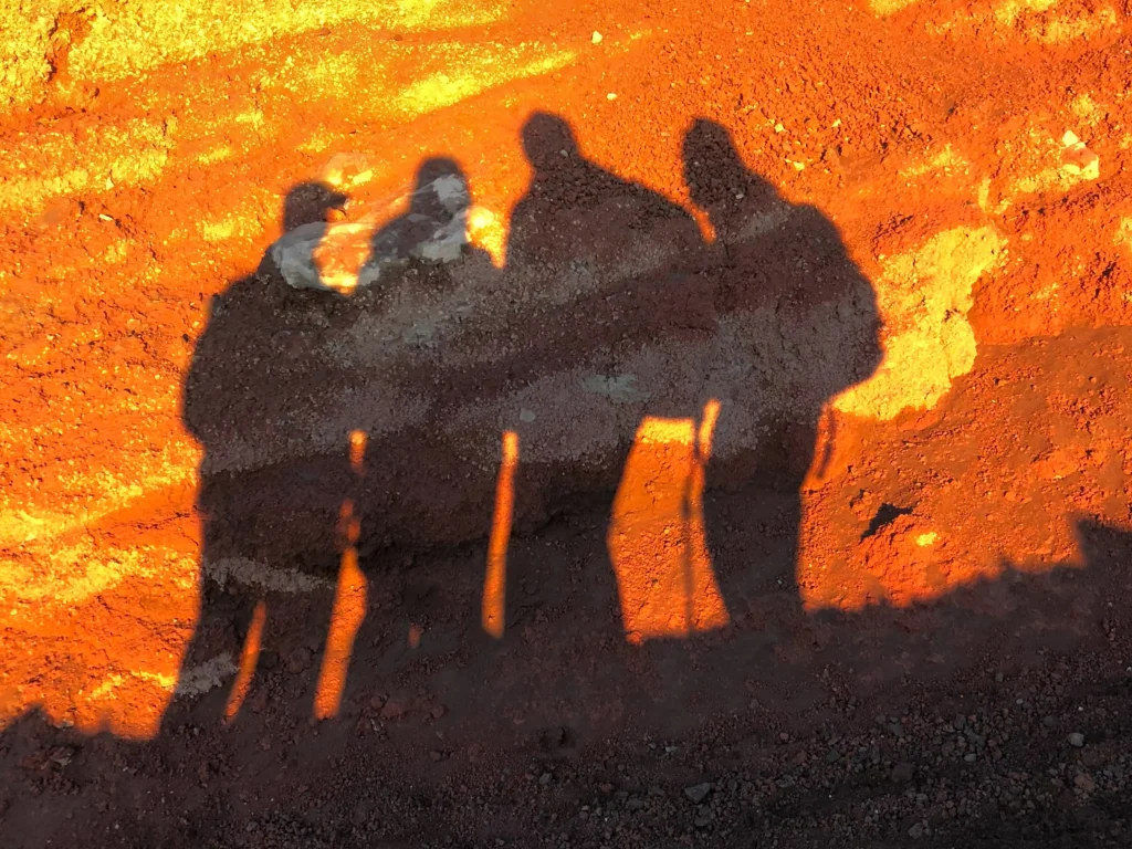 Silhouettes of three climbers cast on orange-red volcanic ground by low morning sun