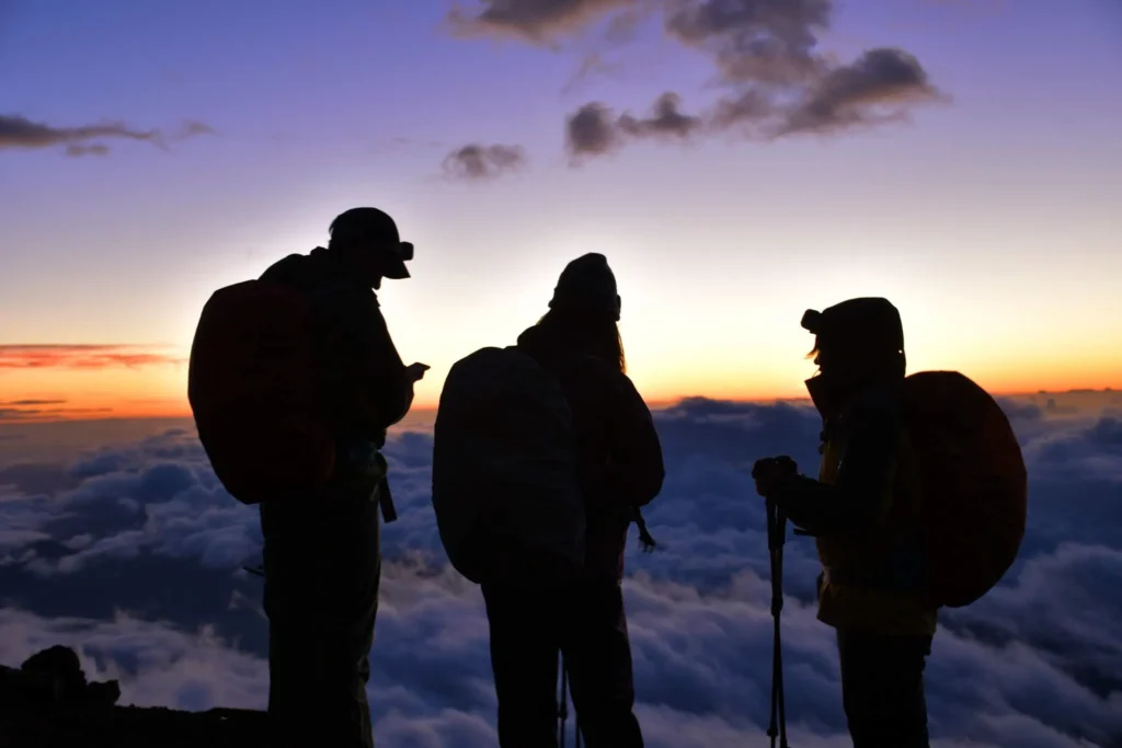 Three climbers silhouetted against a sunset sky, standing above a vast cloud sea