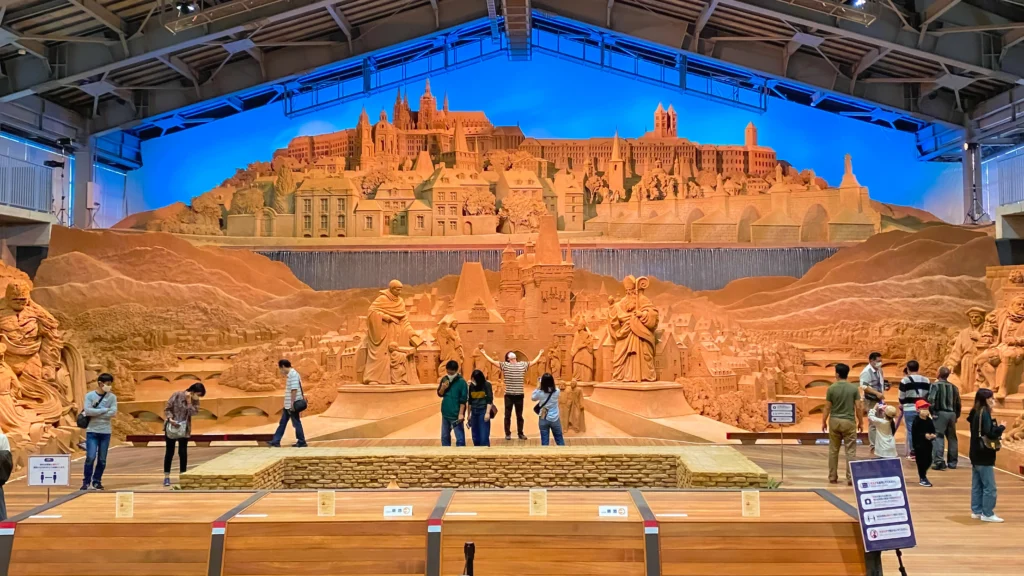 A massive panoramic sand sculpture depicting a European hilltop city with visitors viewing from the floor below