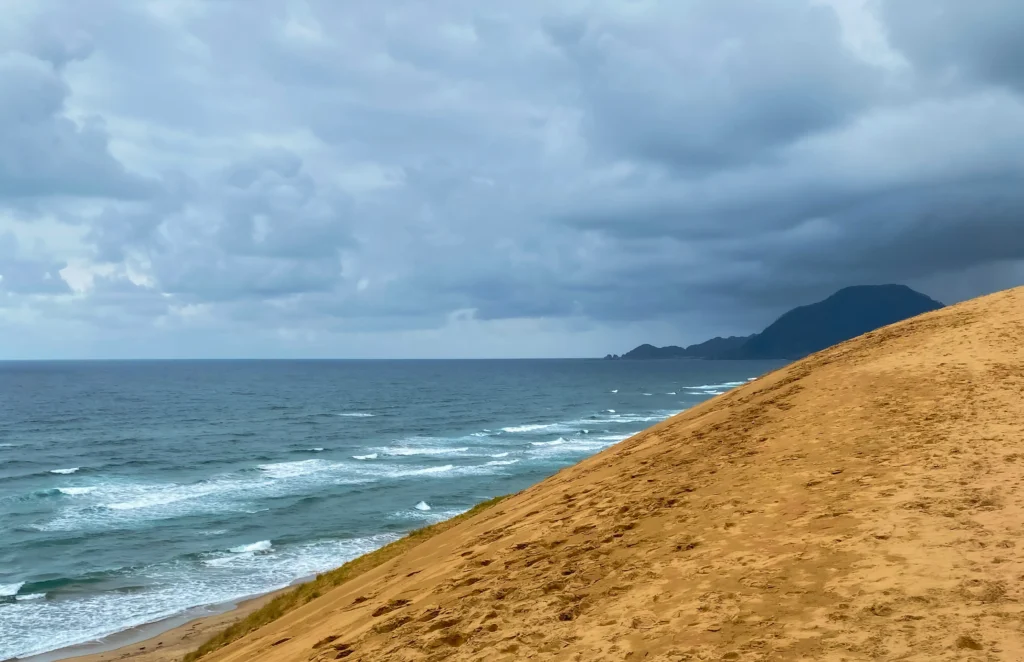 The steep sand slope descending to the Sea of Japan with waves breaking on shore under dramatic clouds